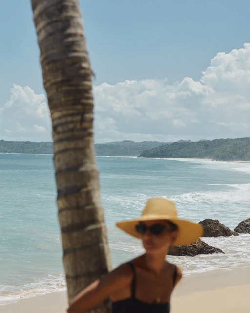Woman in a straw hat stands near a palm tree on a sunny beach with a turquoise sea and distant islands in the background.