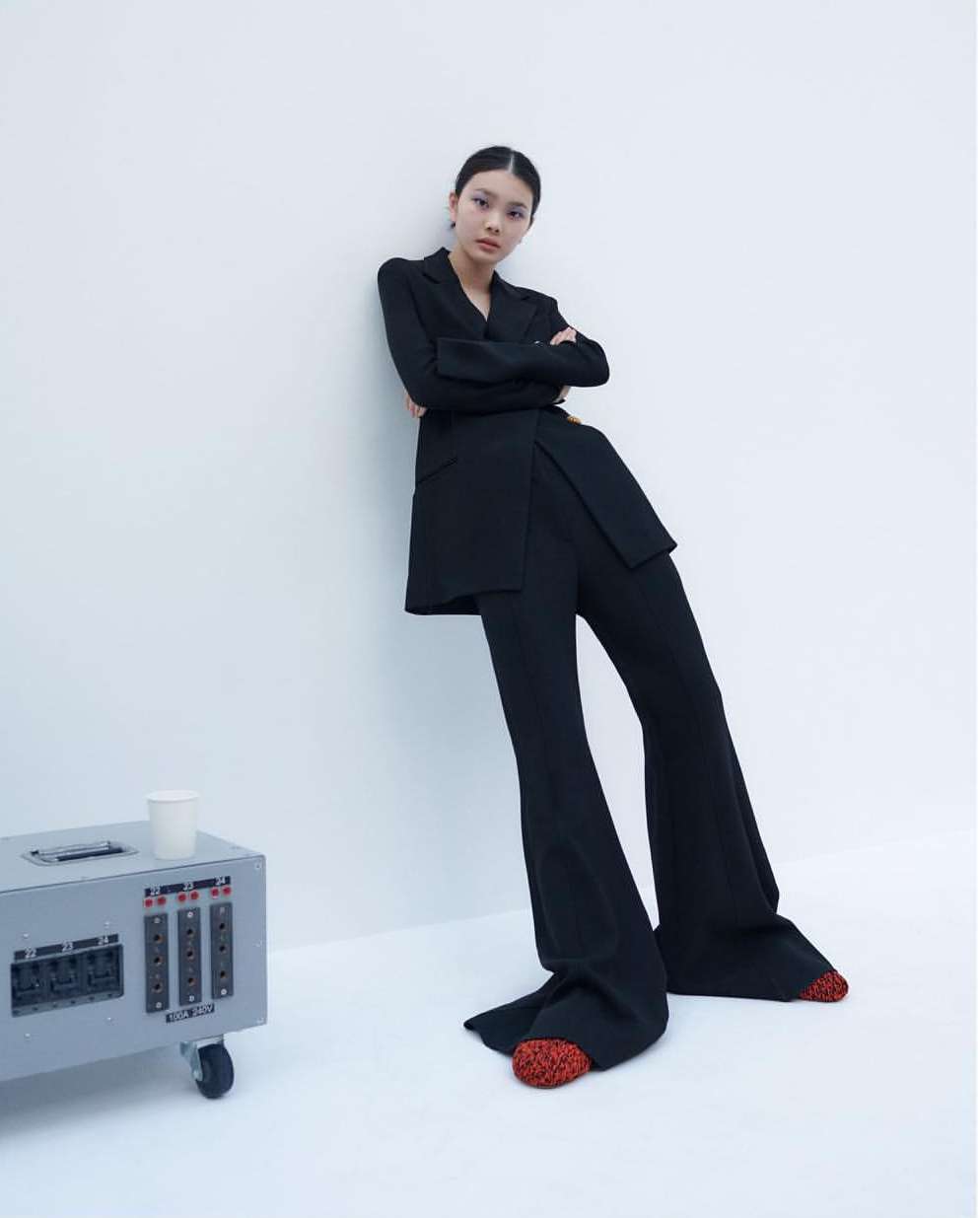 A young woman in a stylish black suit poses against a white wall in a studio, showcasing a modern and elegant fashion aesthetic.