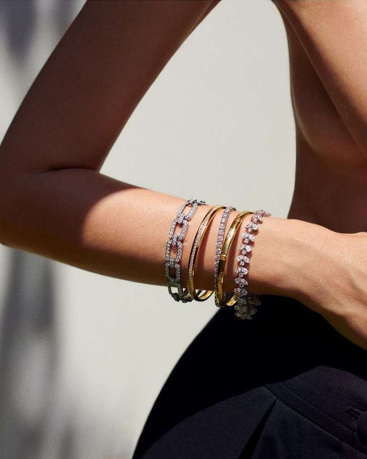A woman poses with an assortment of gold and diamond bracelets, highlighted against a neutral background, showcasing fashion jewelry.