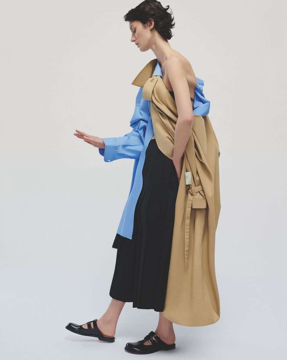 A woman models a blue blouse, black pants, and tan trench coat in a studio, posing in profile against a neutral background.