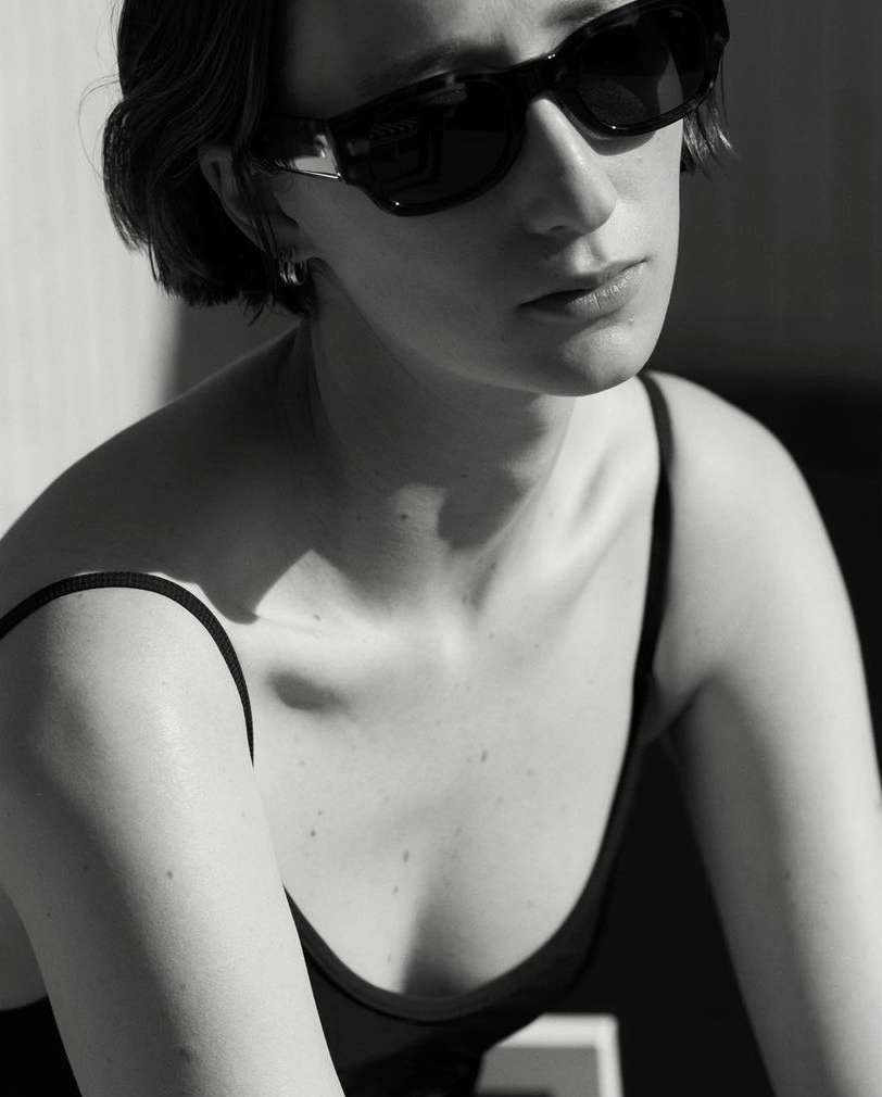 Monochrome portrait of a fair-skinned woman wearing sunglasses and a black top against a light wall in natural, diffused lighting.