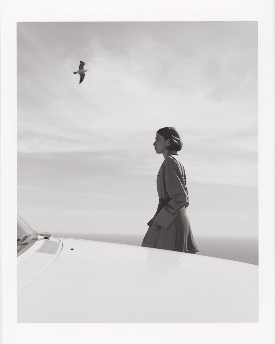 A woman in a coat stands near a vintage car overlooking the ocean, with a seagull flying overhead in a monochrome aesthetic.