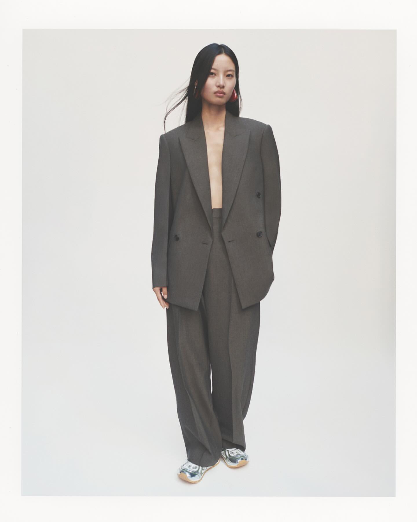 An Asian woman stands in a gray suit with wide-leg pants, accessorized with pink earrings, in a clean studio setting, showcasing fashion.