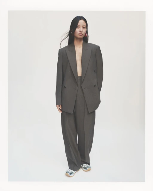 An Asian woman stands in a gray suit with wide-leg pants, accessorized with pink earrings, in a clean studio setting, showcasing fashion.