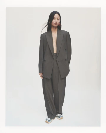 An Asian woman stands in a gray suit with wide-leg pants, accessorized with pink earrings, in a clean studio setting, showcasing fashion.