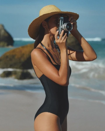Woman in black swimsuit and straw hat holding a vintage camera on a sandy beach