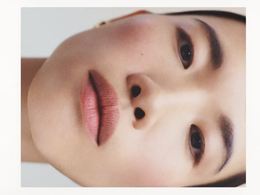 Close-up studio shot of a woman's face featuring soft makeup and neutral lipstick on a light background, capturing beauty and skincare.