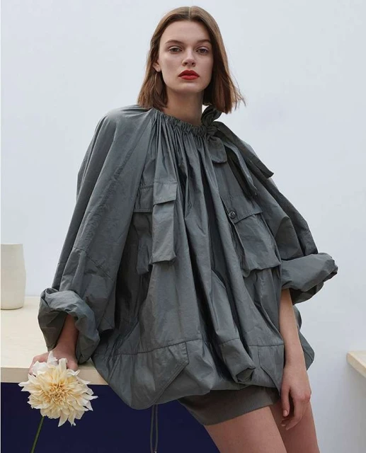 Fashion editorial shot of a female model wearing an oversized grey jacket, neutral shorts, and red lipstick, holding a cream flower.