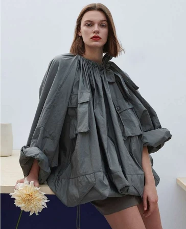 Fashion editorial shot of a female model wearing an oversized grey jacket, neutral shorts, and red lipstick, holding a cream flower.