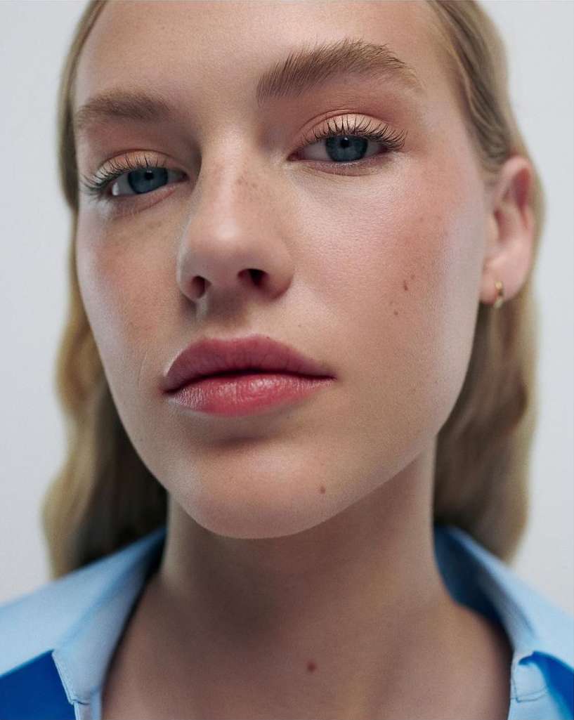 Close-up portrait of a woman with blonde hair, fair skin, and blue eyes, wearing a blue top, against a bright neutral background.