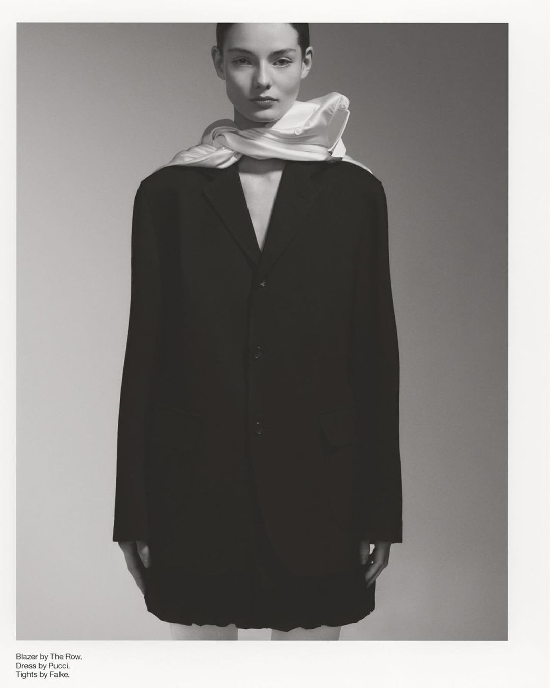 Monochrome fashion portrait of a woman in a black blazer dress with a white scarf, posed against a neutral gray backdrop in studio.