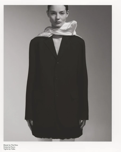 Monochrome fashion portrait of a woman in a black blazer dress with a white scarf, posed against a neutral gray backdrop in studio.