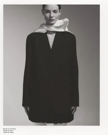 Monochrome fashion portrait of a woman in a black blazer dress with a white scarf, posed against a neutral gray backdrop in studio.
