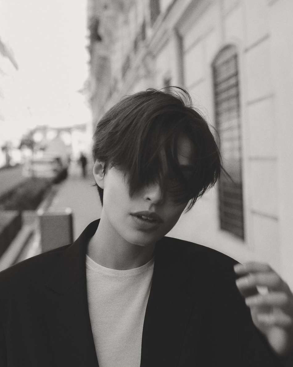Black and white portrait of a young adult with dark hair and a blazer posing outside on a city street with blurred figures.