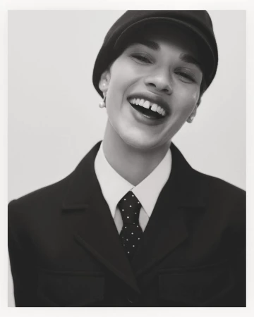 Close-up black and white portrait of a smiling person wearing a dark cap, collared shirt, and polka dot tie.