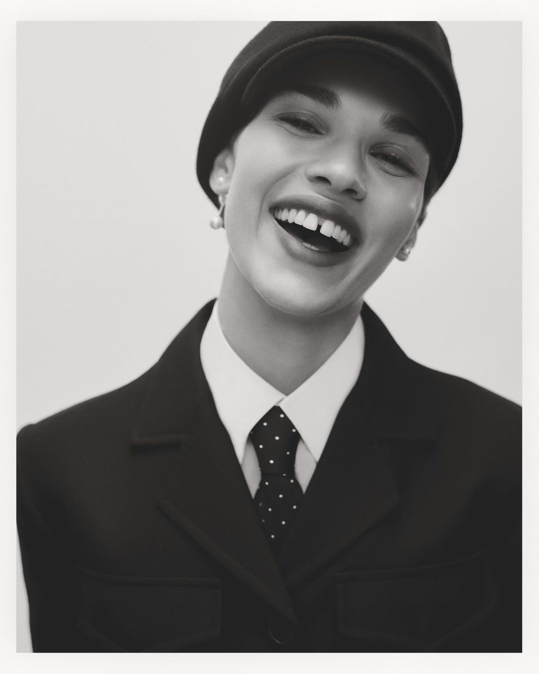 Smiling model wearing a black cap, white collared shirt, and dark jacket against a seamless white background.