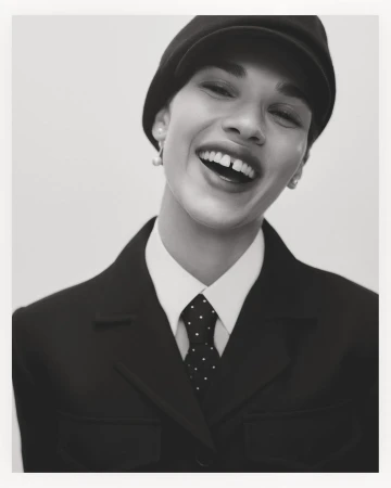 Smiling model wearing a black cap, white collared shirt, and dark jacket against a seamless white background.