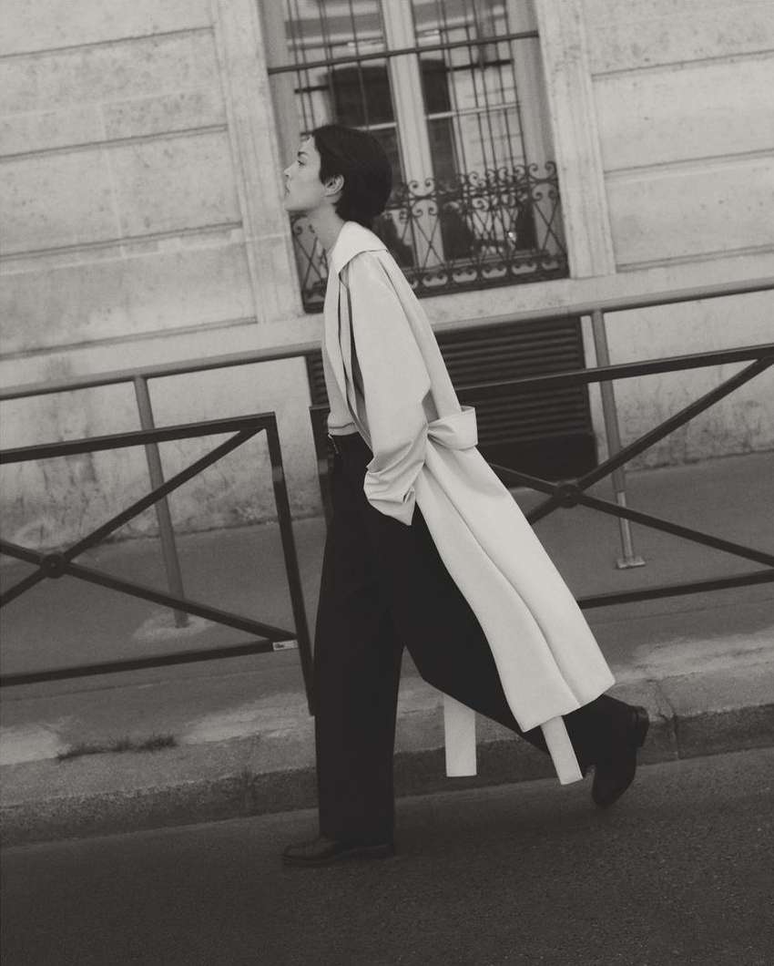 Monochrome outdoor fashion shot of a woman in a trench coat and wide-leg pants walking in an urban setting with a building backdrop.