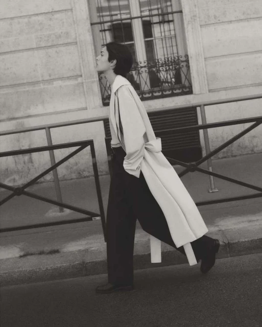 Monochrome outdoor fashion shot of a woman in a trench coat and wide-leg pants walking in an urban setting with a building backdrop.