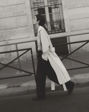 Monochrome outdoor fashion shot of a woman in a trench coat and wide-leg pants walking in an urban setting with a building backdrop.