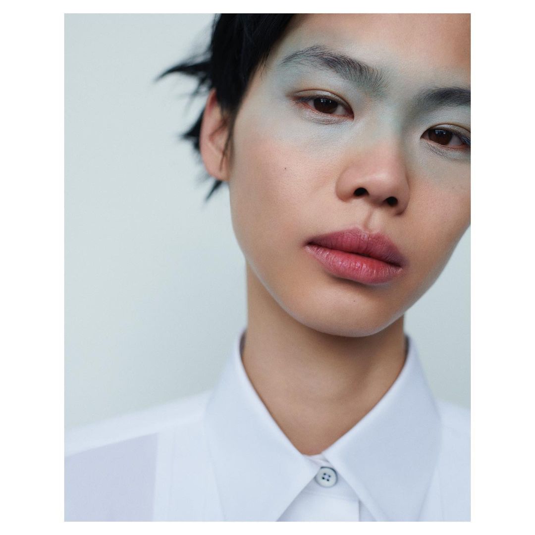 Close-up portrait of a model with pale blue eyeshadow and dark short hair wearing a white button-down shirt.