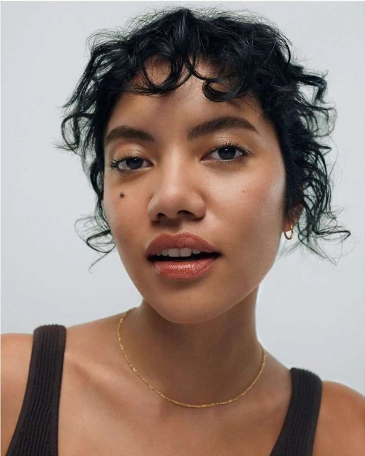 Close-up portrait of a young adult woman with short curly hair and natural makeup, wearing a brown top and a delicate gold necklace.