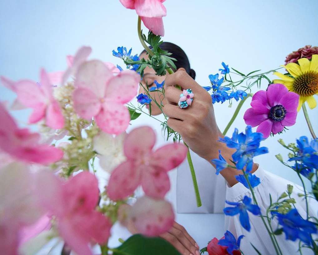 Woman with a ring posing with pink, blue, and yellow flowers against a light blue background, showcasing beauty and elegance.