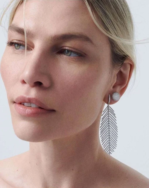 Close-up of a fair-skinned woman with light hair wearing diamond earrings on a neutral background in a vertical format.
