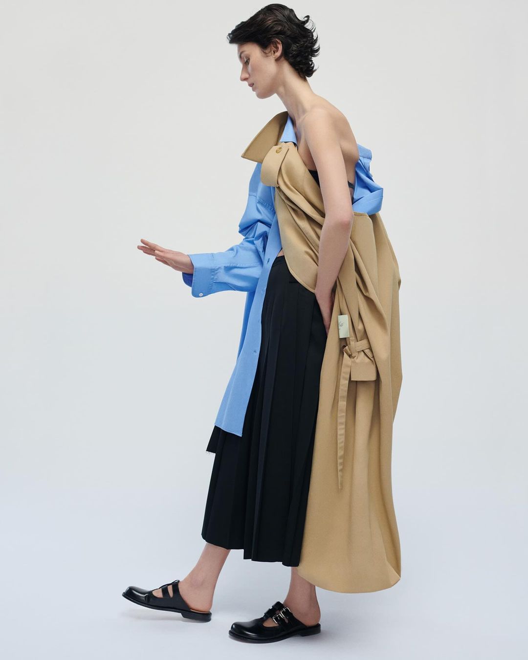 Fashion editorial shot of an adult female model in an open strapless top, midi skirt, dress shirt, coat, and Mary Jane flats in a studio.