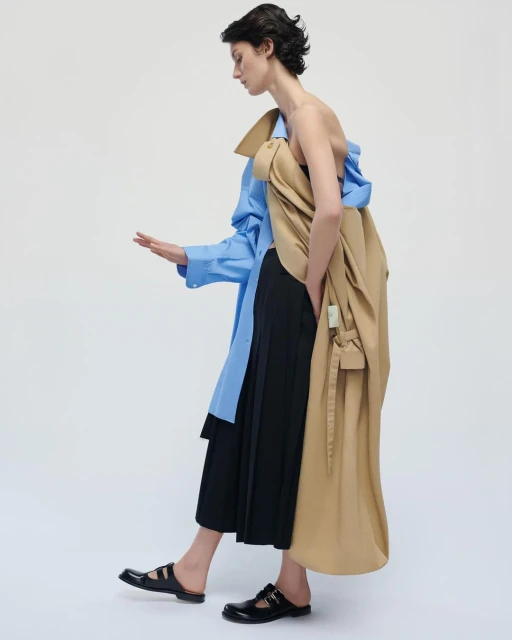 Fashion editorial shot of an adult female model in an open strapless top, midi skirt, dress shirt, coat, and Mary Jane flats in a studio.