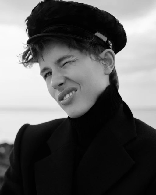 Monochrome portrait of a young adult with a cap, winking and smiling outdoors with water and sky visible in the background.