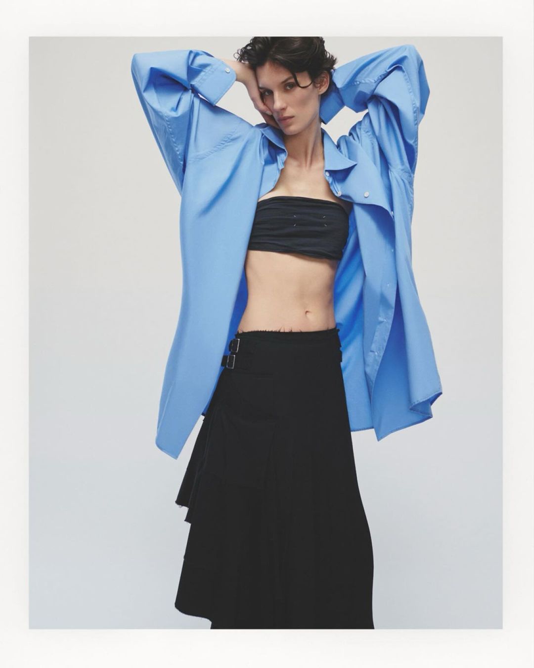Fashionable woman poses in an oversized blue shirt, black tube top, and asymmetrical black skirt in a studio setting with bright lighting.