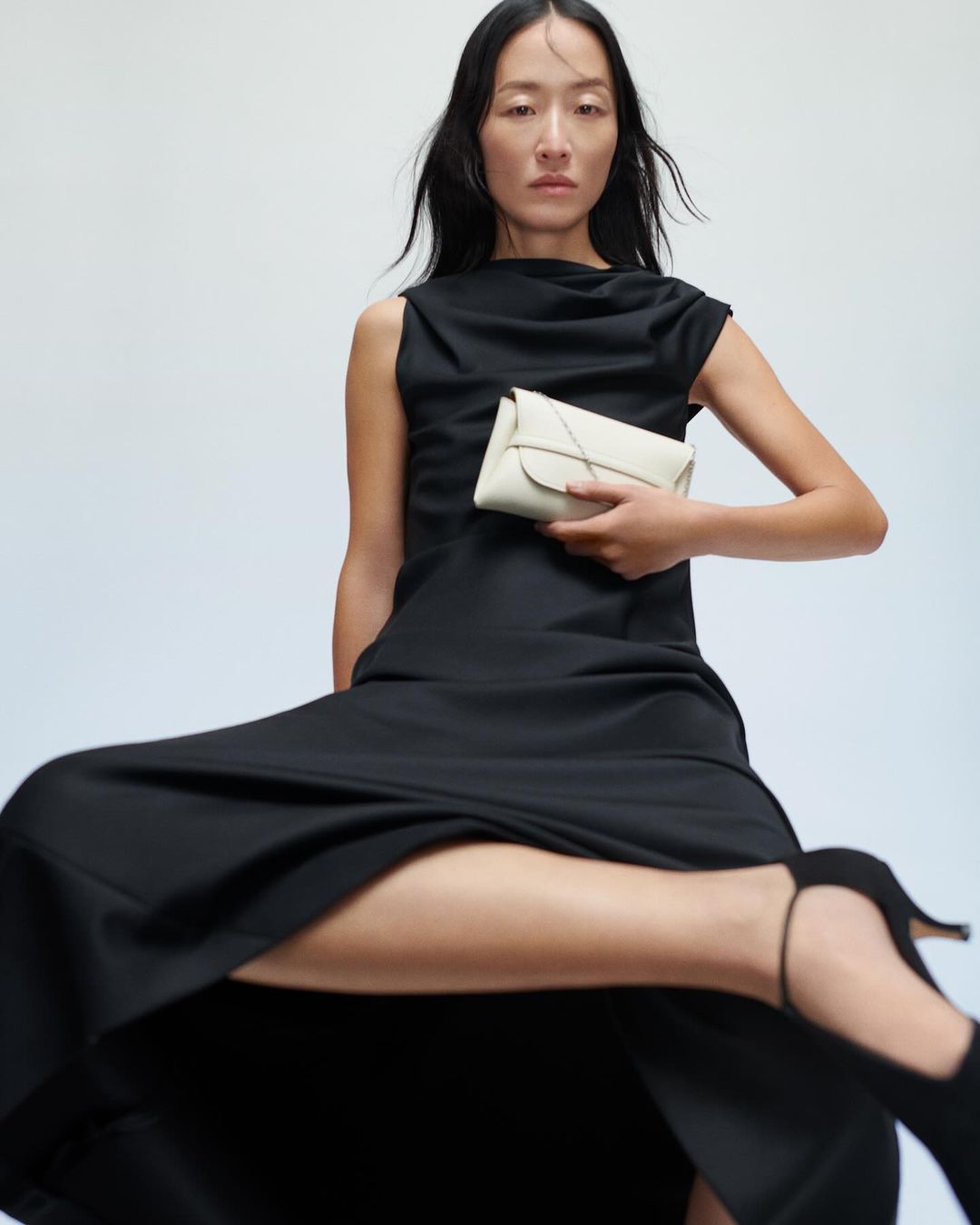 Woman in a long black dress holding a small white handbag against a plain background