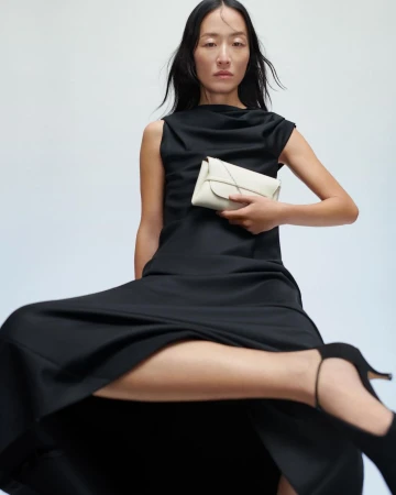 Woman in a long black dress holding a small white handbag against a plain background