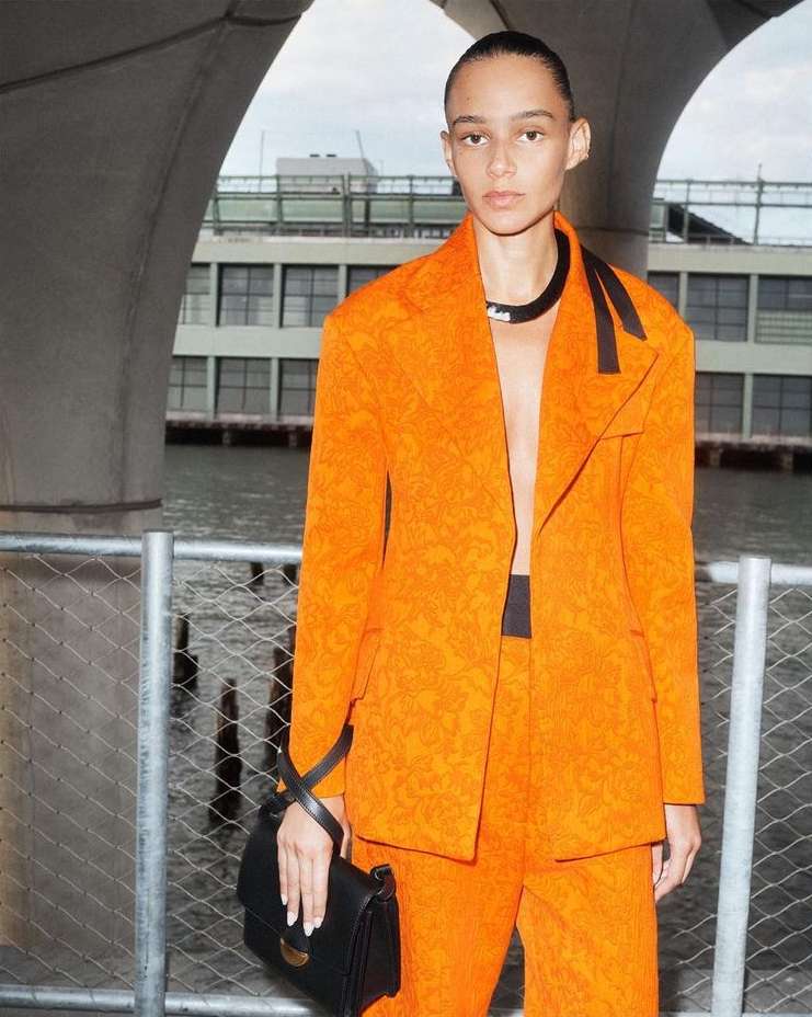 A stylish woman in an orange patterned suit and black choker stands near water, creating a fashionable and vibrant outdoor portrait.