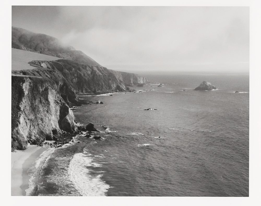 Coastal landscape features a rocky shoreline, cliffs, and ocean waves, creating a dramatic, monochrome scene of natural beauty.