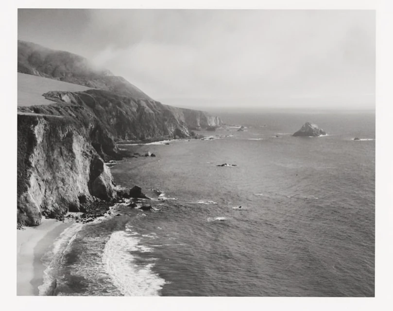 Coastal landscape features a rocky shoreline, cliffs, and ocean waves, creating a dramatic, monochrome scene of natural beauty.