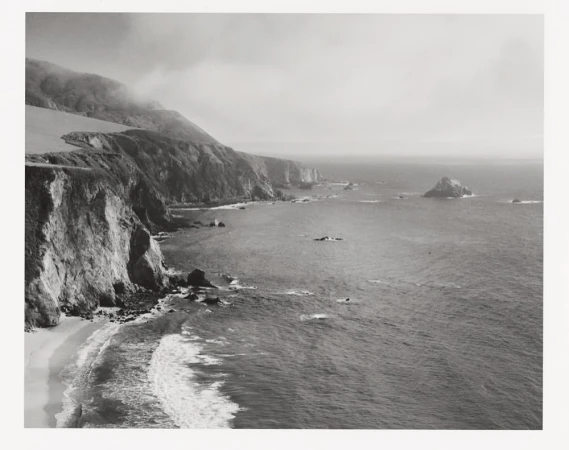 Coastal landscape features a rocky shoreline, cliffs, and ocean waves, creating a dramatic, monochrome scene of natural beauty.