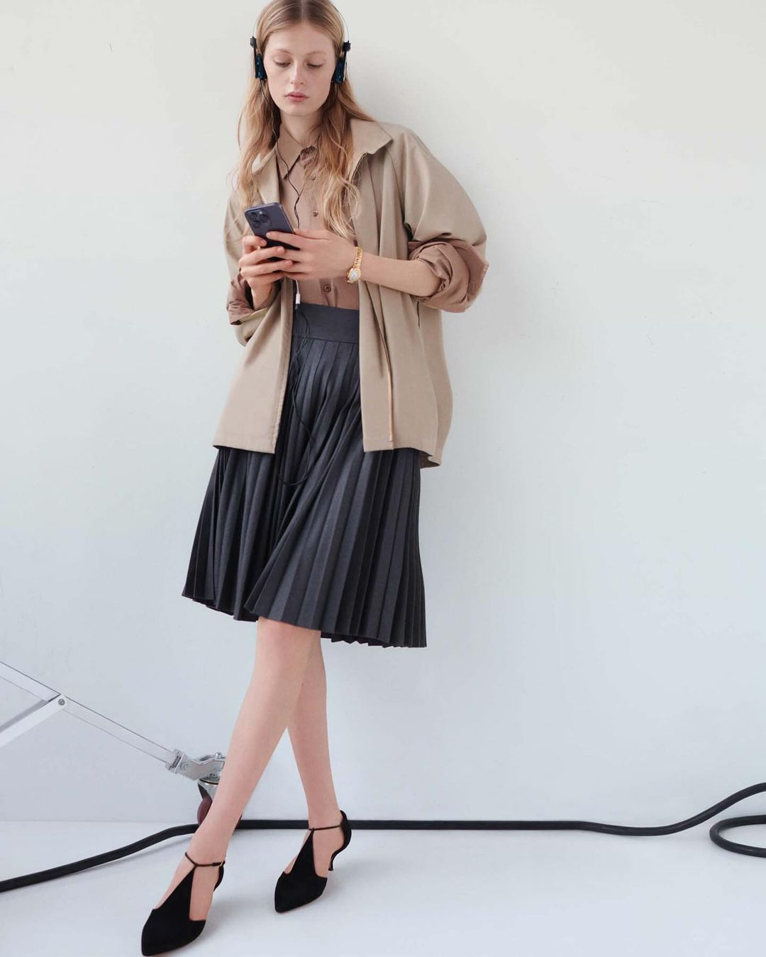 A young woman with headphones looks at her smartphone while wearing a tan jacket, pleated gray skirt, and black t-strap heels.