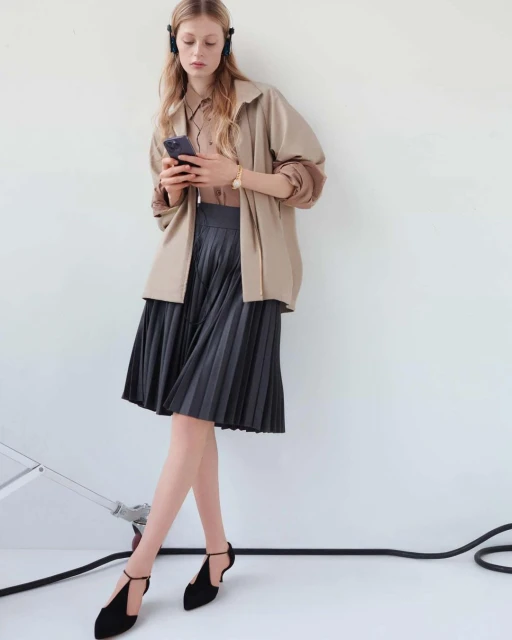 A young woman with headphones looks at her smartphone while wearing a tan jacket, pleated gray skirt, and black t-strap heels.
