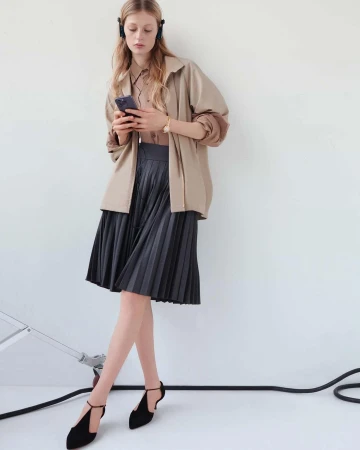 A young woman with headphones looks at her smartphone while wearing a tan jacket, pleated gray skirt, and black t-strap heels.