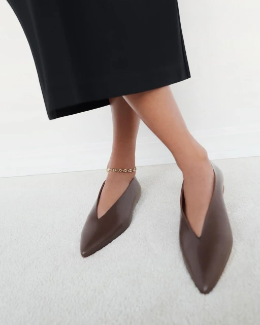 Close-up of woman's legs wearing brown V-cut leather flats and a gold anklet against a white studio backdrop, creating a fashion-forward look.