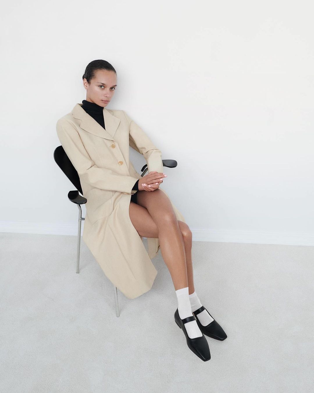 Model seated on a black chair wearing a beige coat, black top, white socks, and black shoes against a plain white wall