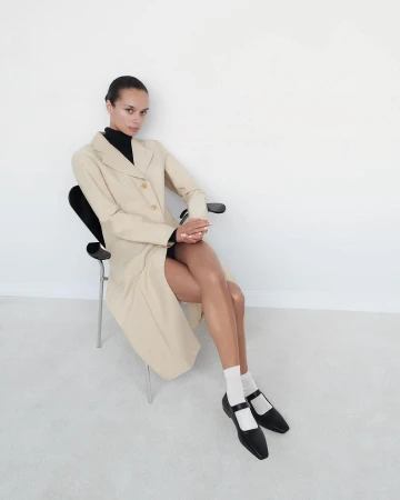 Model seated on a black chair wearing a beige coat, black top, white socks, and black shoes against a plain white wall
