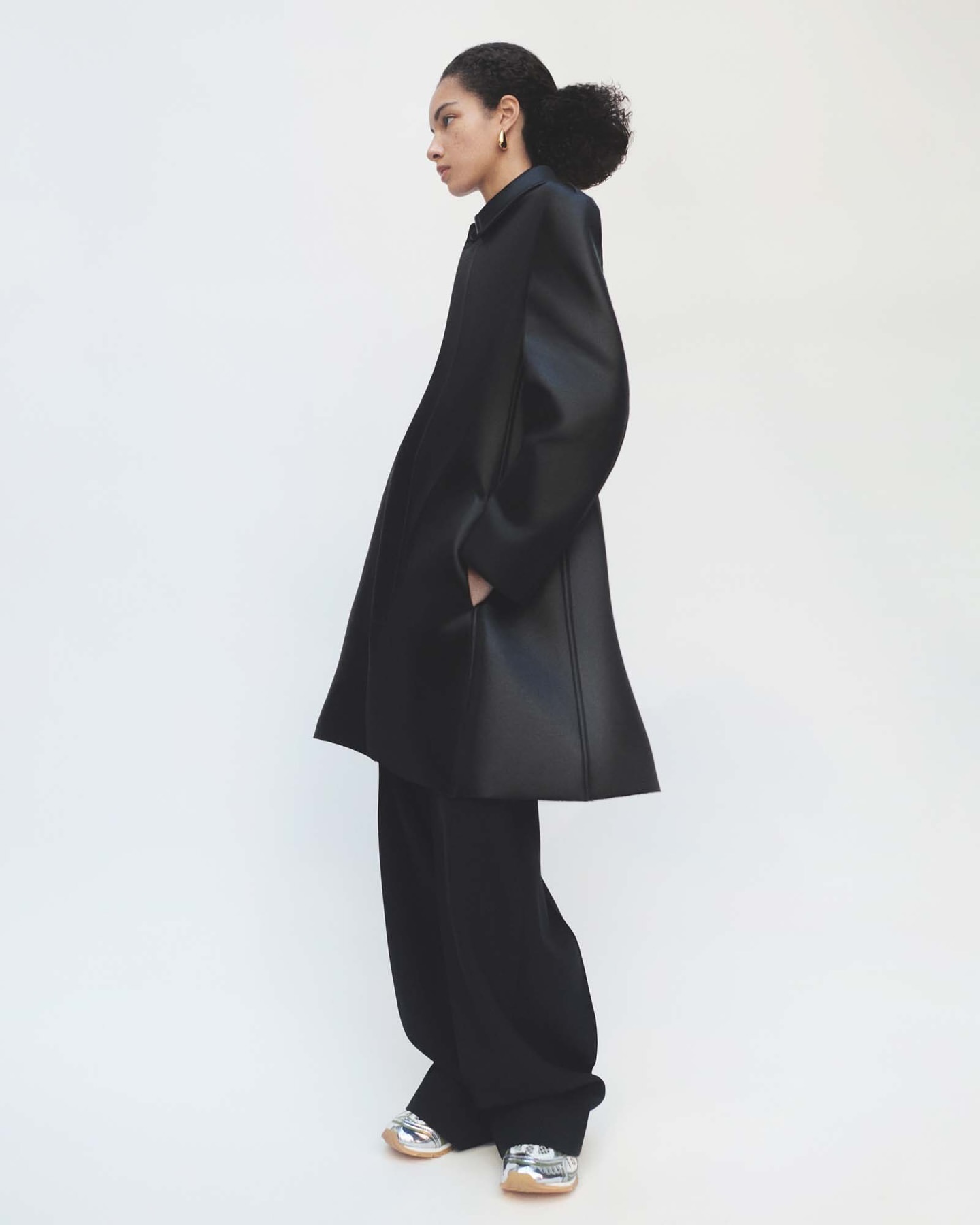 Side profile of an adult woman in a black, oversized coat and wide-legged pants with metallic sneakers, posing in a white photo studio.