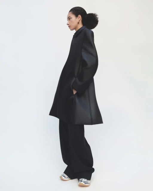 Side profile of an adult woman in a black, oversized coat and wide-legged pants with metallic sneakers, posing in a white photo studio.