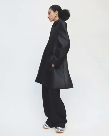 Side profile of an adult woman in a black, oversized coat and wide-legged pants with metallic sneakers, posing in a white photo studio.