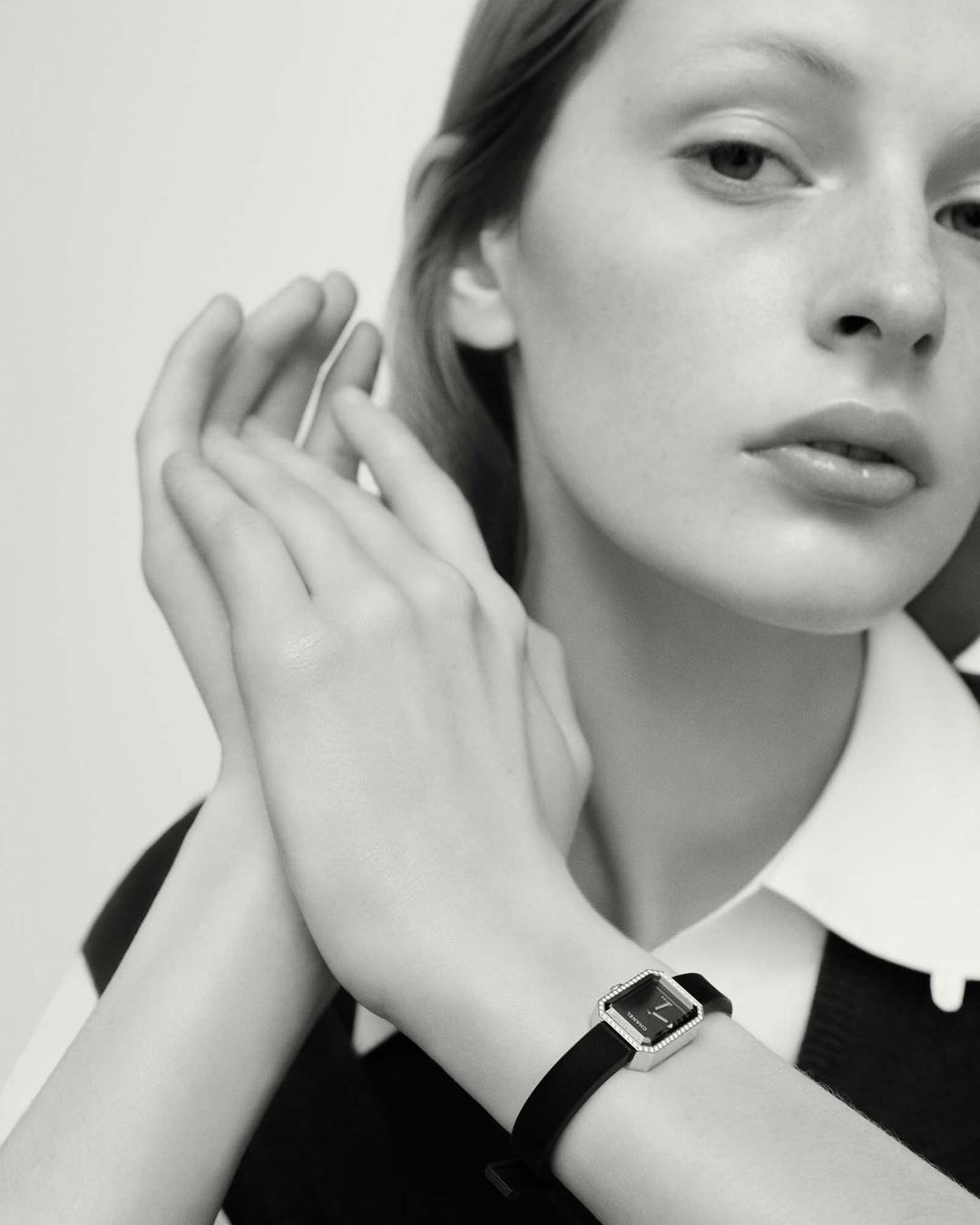 Monochrome close-up of a fair-skinned woman wearing a square-faced wristwatch with a black band in a brightly lit indoor setting.