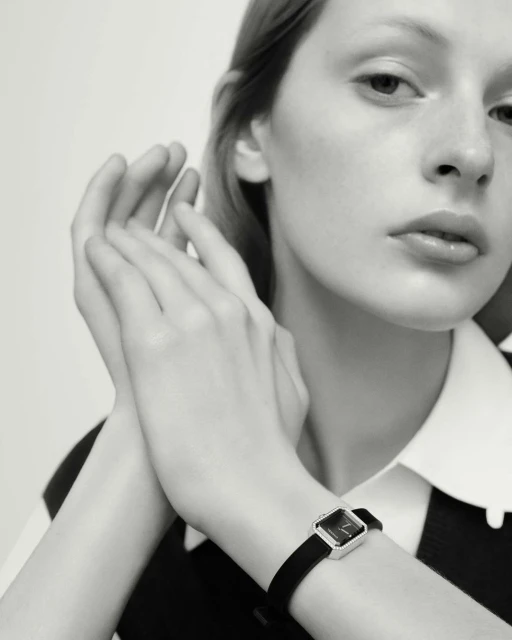 Monochrome close-up of a fair-skinned woman wearing a square-faced wristwatch with a black band in a brightly lit indoor setting.