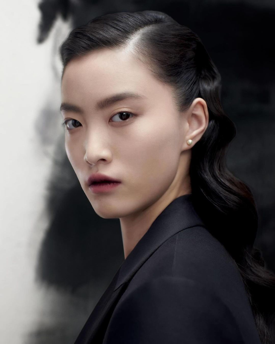 Close-up portrait of a young Asian woman with dark hair, wearing a black blazer against a soft, blurred black and white backdrop.